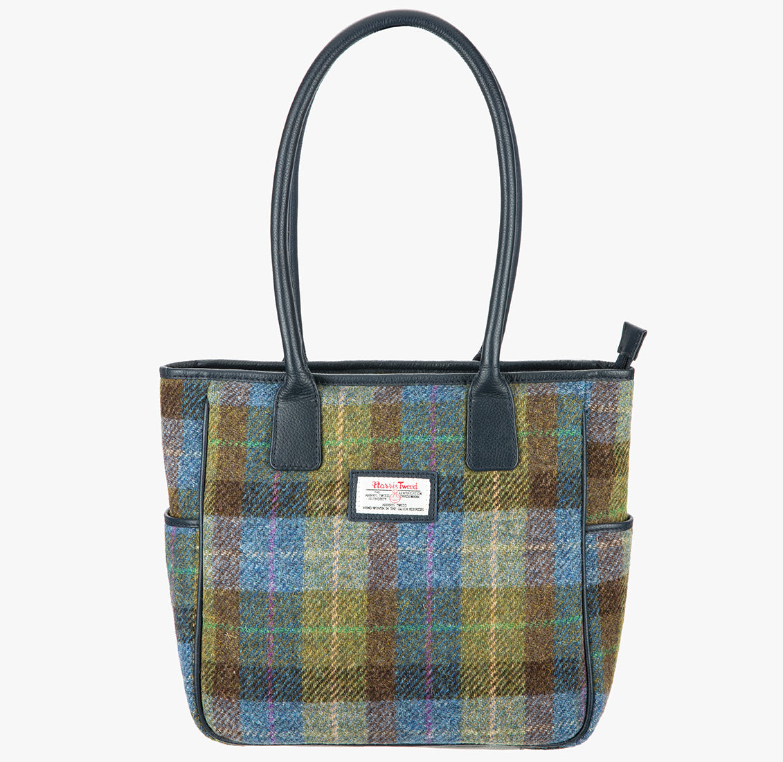 Harris Tweed tote bag in a blue, green and brown check with navy blue leather handles that go over the shoulder. It has a pocket on each side of the bag trimmed with navy blue leather. It also has a harris tweed logo on the front of the bag in the middle which is sewn onto a leather label holder.
