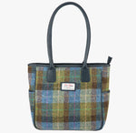 Harris Tweed tote bag in a blue, green and brown check with navy blue leather handles that go over the shoulder. It has a pocket on each side of the bag trimmed with navy blue leather. It also has a harris tweed logo on the front of the bag in the middle which is sewn onto a leather label holder.