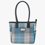 Harris Tweed tote bag in a pale blue, purple and grey check with navy blue leather handles that go over the shoulder. It has a pocket on each side of the bag trimmed with navy blue leather. It also has a harris tweed logo on the front of the bag in the middle which is sewn onto a leather label holder.