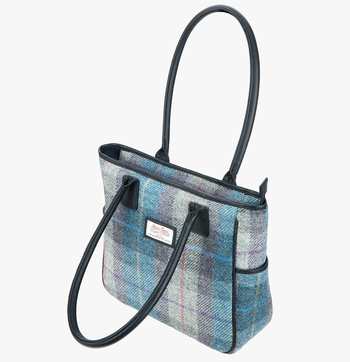 Harris Tweed tote bag with handles that go over the shoulder, it is a pale blue, purple, and grey check. This bag is Lavender Harris Tweed with navy blue leather handles. It has a pocket on each side of the bag trimmed with navy leather. This bag has the front folded over.