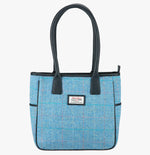 Harris Tweed tote bag in sky with black leather handles that go over the shoulder.  It has a pocket on each side of the bag trimmed with black leather. It also has a harris tweed logo on the front of the bag in the middle which is sewn onto a leather label holder.