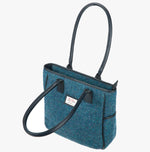 Harris Tweed tote bag with handles that go over the shoulder. This bag is teal harris tweed with black leather handles. It has a pocket on each side of the bag and a harris tweed logo on the front of the bag. It is shown with the front handle folded over.