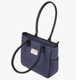 Harris Tweed tote bag with handles that go over the shoulder. This is shown with the front handle folded forward. This bag is violet Harris Tweed with black leather handles. It has a pocket on each side of the bag trimmed in black leather and a harris tweed logo on the front in the middle of the bag.
