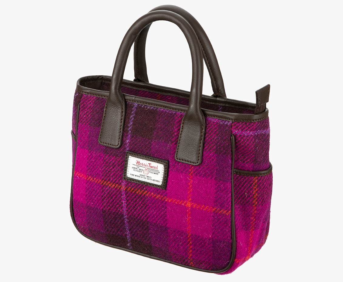 This is a three-quarter view of the Harris Tweed handheld bag. This bag's colour is bright pink and dark pink check. It has a pocket on each side of the bag trimmed with brown leather. It also has short brown leather handles.