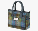 This is a three-quarters view of the Harris Tweed handheld bag. The bag's colour is blue, green and brown check. It has a pocket on each side of the bag trimmed with navy leather. It also has short navy blue leather handles.