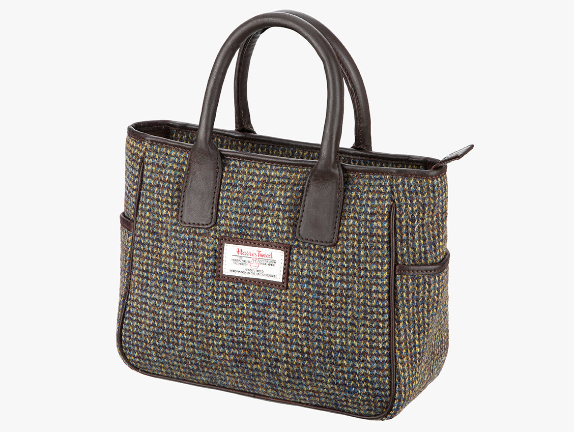 This is a three-quarters view of the Harris Tweed handheld bag. The bag's colour is heath which is a mustard, brown and blue design. It has a pocket on each side of the bag trimmed with brown leather. It also has short brown leather handles.