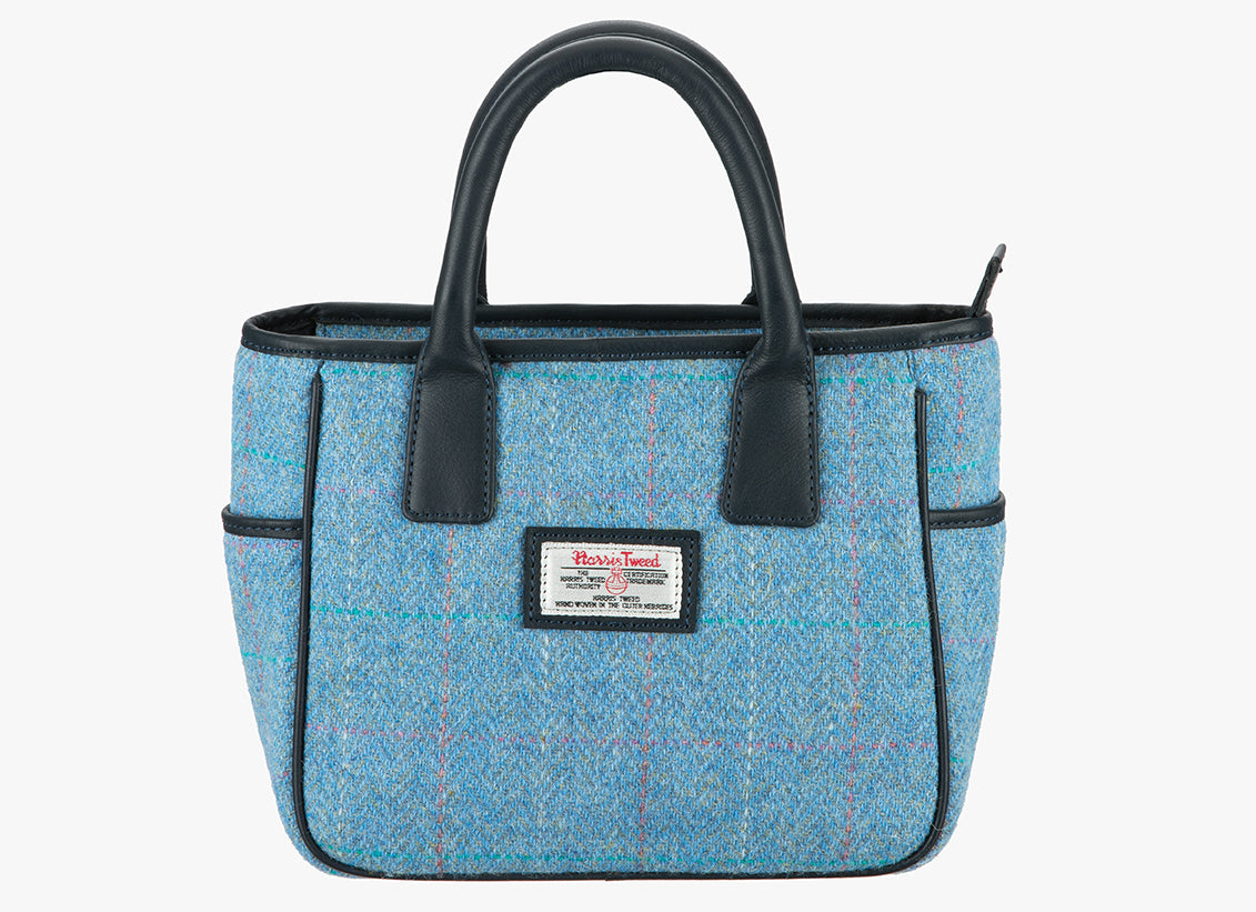 Harris Tweed handheld bag in sky with navy leather handles. The colour is a sky blue with very fine  pink, turquoise and white overcheck . It has a pocket on each side of the bag trimmed with navy leather. It also has a Harris Tweed logo on the front of the bag in the middle which is sewn onto a leather label holder.