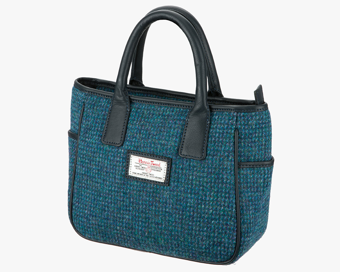 This is a three-quarter view of the Harris Tweed handheld bag. This bag's colour is teal. It has a pocket on each side of the bag trimmed with black leather. It also has short black leather handles.