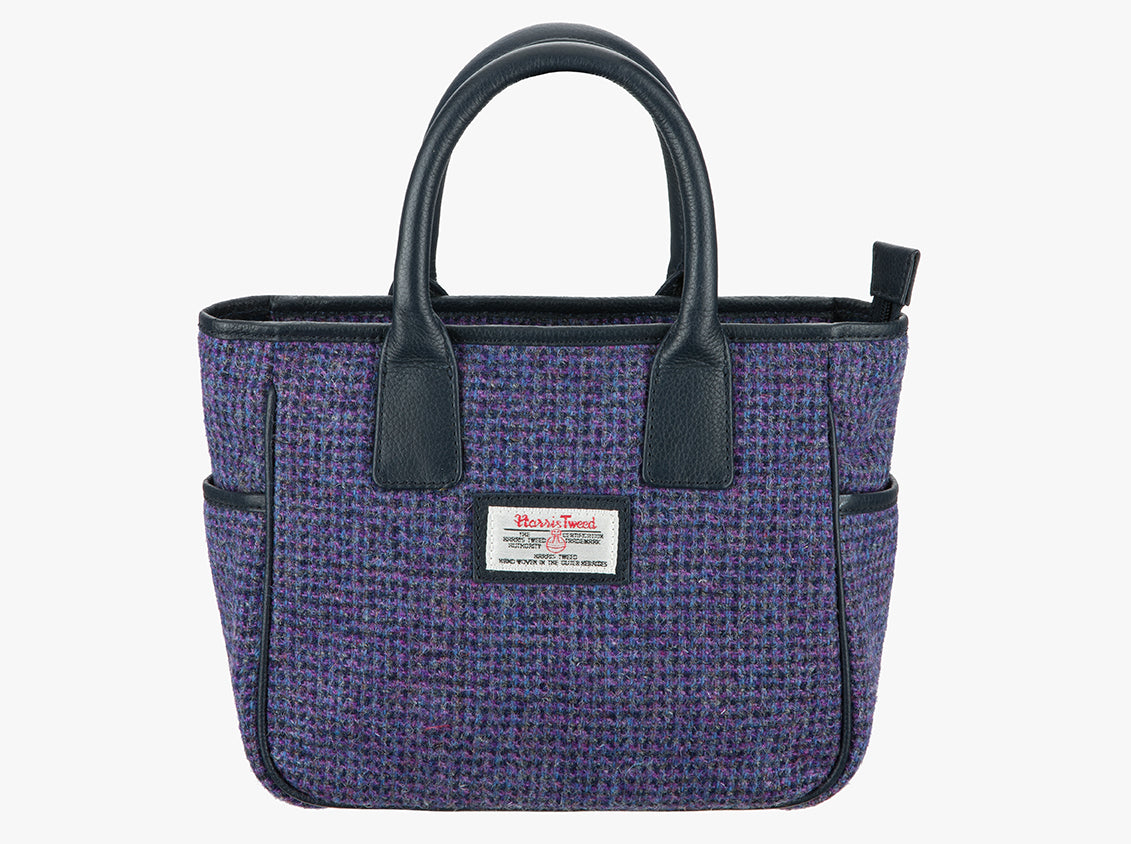 Harris Tweed handheld bag in a violet check with black leather handles. The colour is a violet check which is a purple and black fine check . It has a pocket on each side of the bag trimmed with black leather. It also has a Harris Tweed logo on the front of the bag in the middle which is sewn onto a leather label holder.