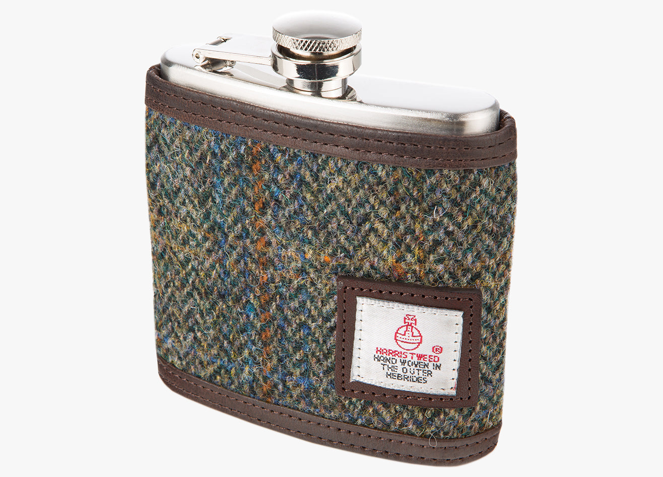 Harris Tweed Hip flask in green and blue and brown. The cover is Harris tweed and leather. The hip flask holds six ounces and is made from stainless steel. The Harris tweed is green, blue and brown herringbone, it has brown leather at top and bottom of the cover. It also has a small square Harris Tweed logo on the bottom right.