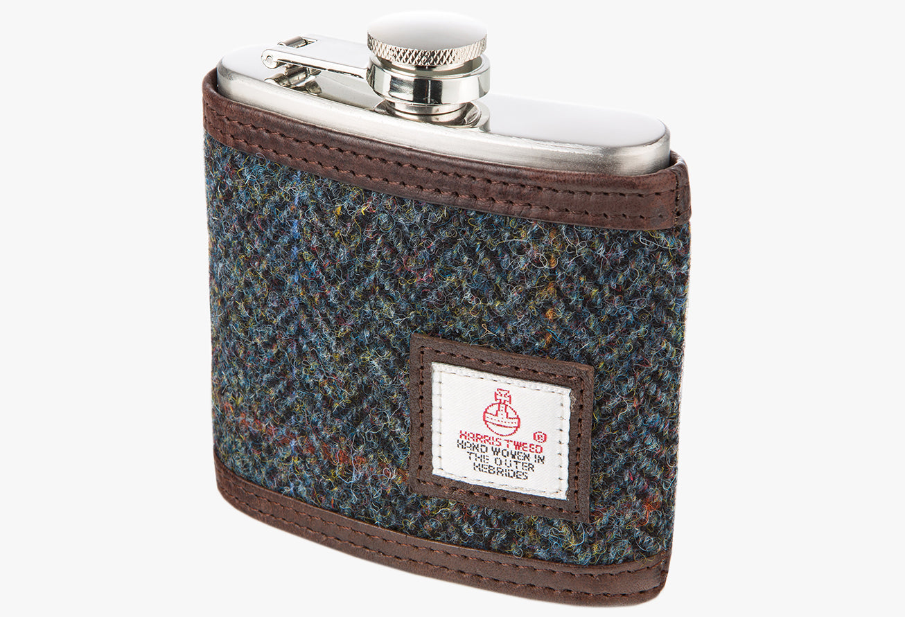 Harris Tweed Hip flask in midnight Blue. The cover is Harris tweed and leather. The hip flask holds six ounces and is made from stainless steel. The Harris tweed is navy blue with a fine overcheck in rust brown, it also has brown leather at top and bottom of the cover. It also has a small square Harris Tweed logo on the bottom right.