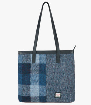 Harris Tweed shopper with navy blue leather handles that sit comfortably on the shoulder. It also has navy blue leather trim at the top of the bag. The bag design is in two halves; the left half is a blue check Harris Tweed which is navy, mid-blue and light blue check. The right side is a light blue and dark blue herringbone design. It also has a small rectangular Harris Tweed logo at the bottom right.