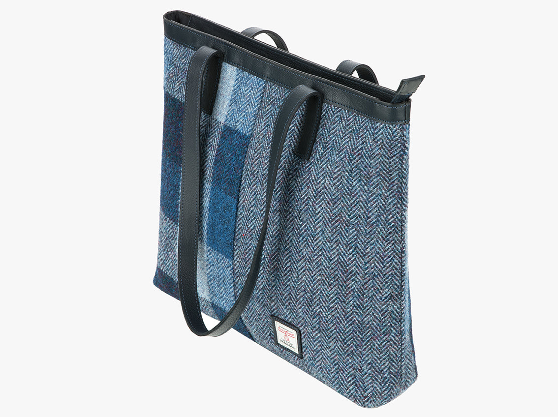 Three-quarters view of the Harris Tweed shopper with a navy blue leather handle, which is shown folded over however it can sit comfortably on the shoulder. It also has a navy blue leather trim at the top of the bag. The bag design is in two halves, the left half is a blue Harris Tweed check, this is navy, light blue and mid-blue check. The right side is a small check in purple and black. It also has a small square harris tweed logo at the bottom right.