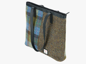 Three-quarters view of the Harris Tweed shopper with a  navy blue leather handle which is shown folded over however it can sit on the shoulder. It also has a navy blue leather trim at the top of the bag. The bag design is in two halves, the left half is denim sage harris tweed, this is green and blue check, the right side is heath which is a brown and mustard small herringbone design. It also has a small square harris tweed logo at the bottom right.