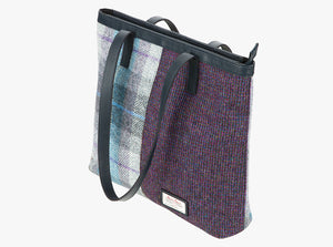 Three-quarters view of the Harris Tweed shopper with a navy blue leather handle which is shown folded over however it can sit on the shoulder. It also has a navy blue leather trim at the top of the bag. The bag design is in two halves, the left half is Lavender Harris Tweed which is grey, lilac, and blue check. The right side is a small check in purple and black. It also has a small square harris tweed logo at the bottom right.