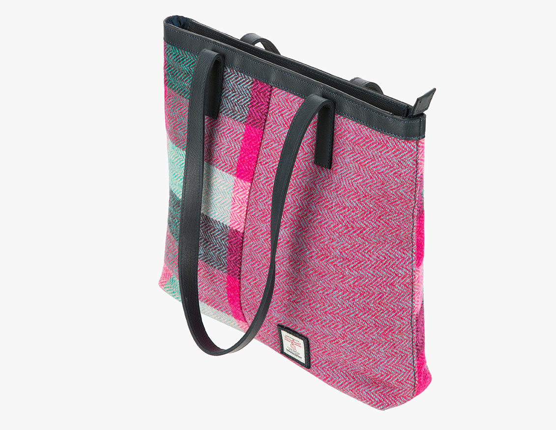 Three-quarters view of the Harris Tweed shopper with black leather handles which are shown folded over however, they can sit on the shoulder. It also has black leather trim at the top of the bag. The bag design is in two halves, the left half is pink Harris Tweed which is bright pink, jade green, and light green check. The right side is a bright pink and pale blue herringbone. It also has a small square harris tweed logo at the bottom right.